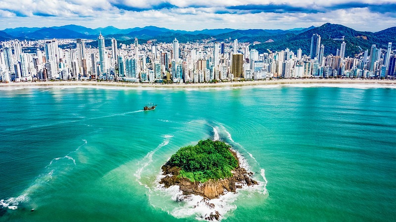 Imagem panorâmica de Balneário Camboriú, mostrando  uma ilha centralizada, cercada por água azul esverdeada e, ao fundo, a linha da praia com seus prédios brancos.