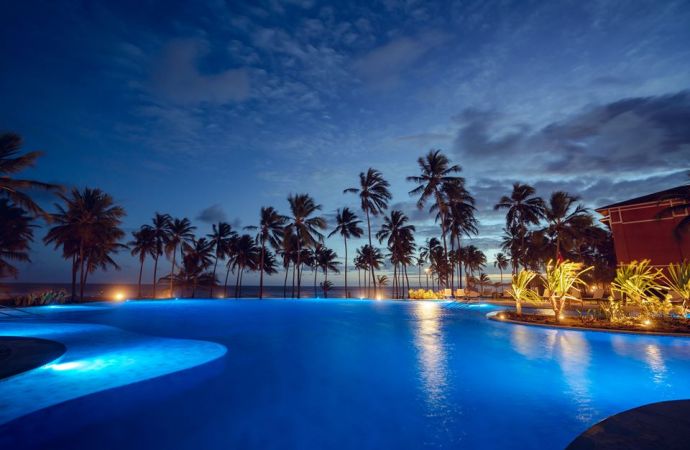 Piscina iluminada por holofotes cercada por palmeiras sob um céu já anoitecendo