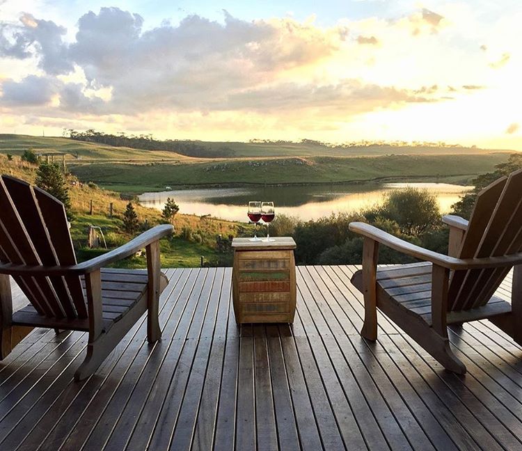 Duas poltronas confortáveis sore um deck de madeira junto a taças de vinho cheias de frente para uma paisagem campestre com árvores e um lado sob o céu da tarde em hotéis e resorts em santa catarina