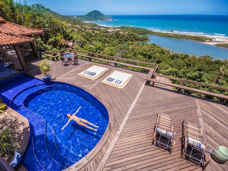 Imagem vista de cima, um deck de madeira, com piscina e uma moça dentro dela, ao fundo, a natureza e um azul da uma praia