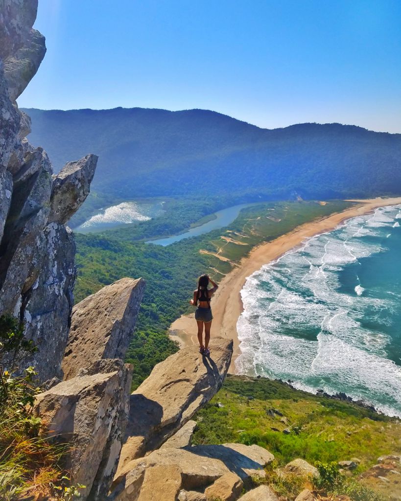 Visão de um dos mirantes da praia da Lagoinha do Leste, com montanhas rochosas, cobertas pela vegetação e uma faixa de areia comprida a frente de um mar azul agitado.