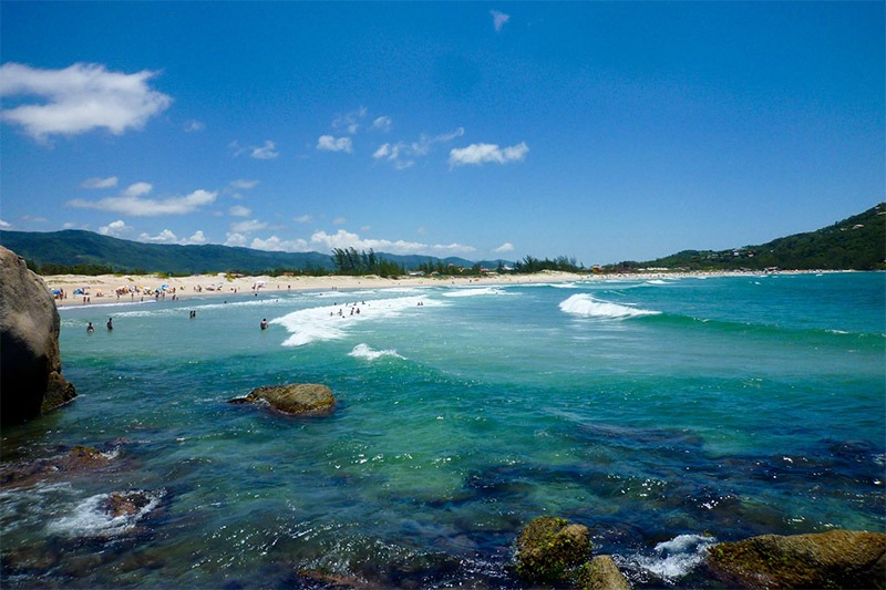 quantas praias tem em Santa Catarina? Foto da praia da Ferrugem, com seu mar azula esverdeado, algumas rochas, a faixa de área com algumas pessoas e a natureza a sua volta