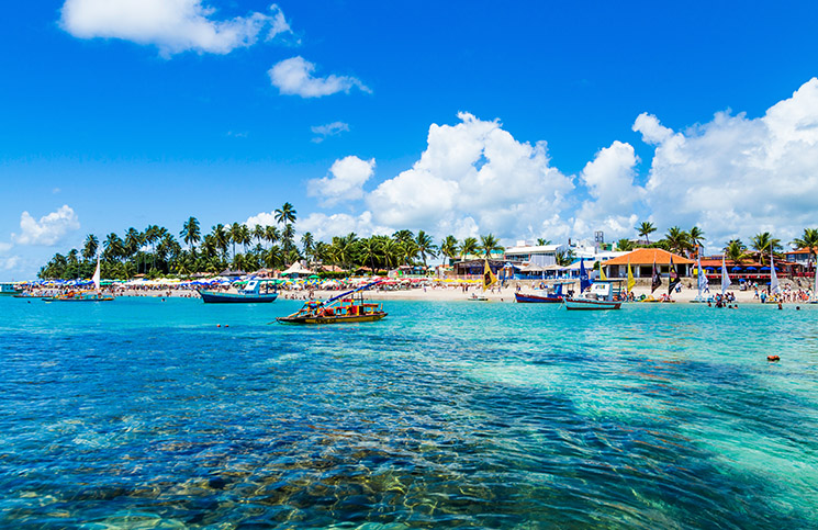 Vista do mar esverdeado de Porto de Galinhas com barquinhos coloridos, praia cheia de pessoas, natureza e alguns prédios em hotel em porto de galinhas beira mar