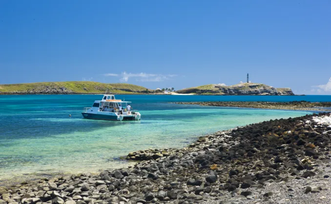 Rochas, mar azulado, barco turístico e ao fundo, a ilha de Santa Bárbara com seu farol sob o céu azul.