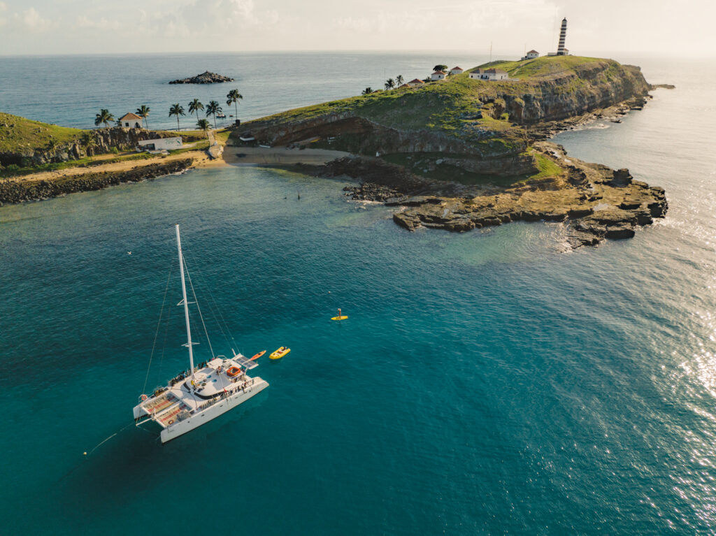 Imagem vista de cima de um catamarã sobre a o mar azulado e a ilha de Santa Bárbara com casas, plameiras e o farol em Abrolhos Bahia 