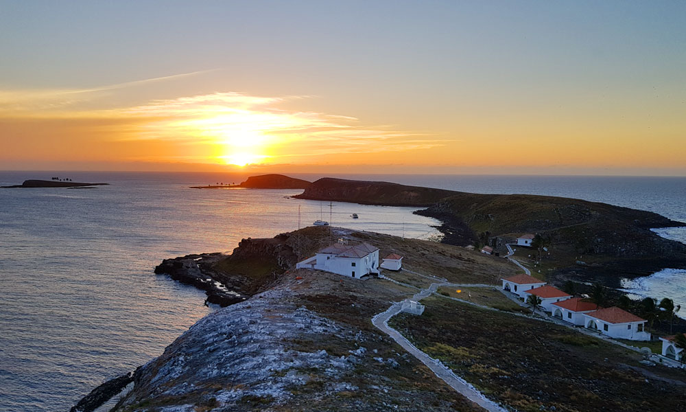 Imagem do alto da ilha de Santa Bárbara, com casas e uma estradinha sob o pôr do sol.