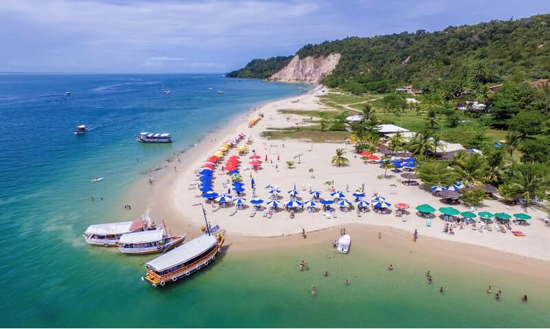 Barcos parados em uma praia de águas claras e verdejantes a frente de barraquinhas na areia