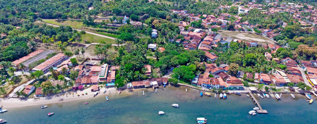 Foto panorâmica com vários prédios e casas entre uma pequena floresta e o mar azul em onde fica Boipeba na Bahia