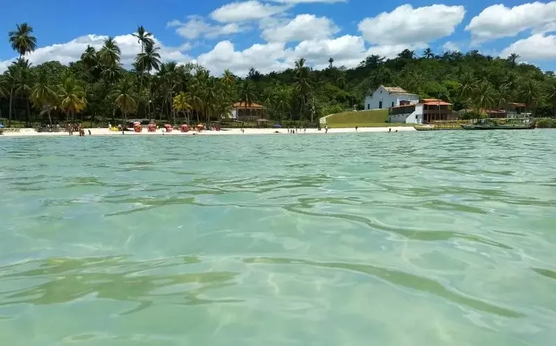 Imagem do nível da água da praia de praia das neves bahia, mostrando água cristalina, areia,  palmeiras e coqueiros e a igreja de Nossa Senhora das Neves