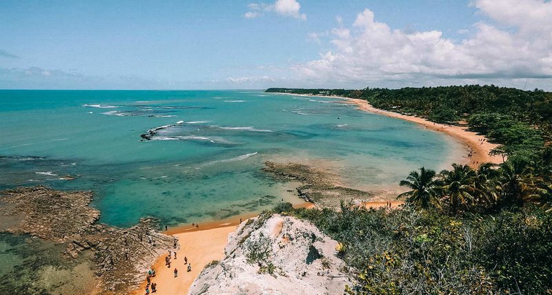 Imagem vista de cima de uma falésia coberta por vegetação nativa com a praia de areia branca embaixo e o mar azulado até o horizonte de Trancoso praia do Espelho